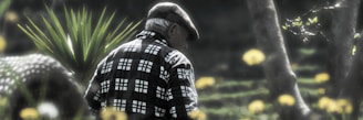 An elderly person wearing a checkered shirt and a flat cap walks through a garden or park setting. The background is filled with blurred yellow flowers and greenery, creating a serene and nostalgic atmosphere.