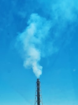 A tall industrial chimney emits thick white smoke against a clear blue sky. The smoke forms a large plume, dispersing as it rises higher into the atmosphere.