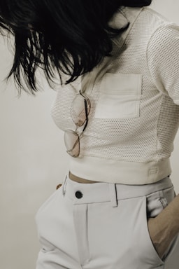 A person wearing a white mesh top with a front pocket and high-waisted white trousers. Sunglasses hang from the pocket, and the person has dark, shoulder-length hair.