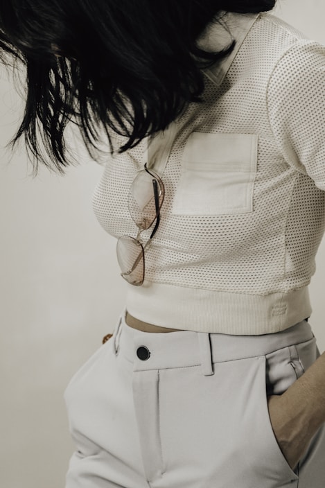 A person wearing a white mesh top with a front pocket and high-waisted white trousers. Sunglasses hang from the pocket, and the person has dark, shoulder-length hair.