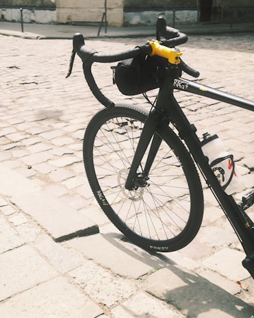 A bicycle with a black frame and yellow handle stem is resting on cobblestone pavement. The bicycle has a black handlebar with a small bag attached, and a visible water bottle mounted on the frame. The front wheel is prominently displayed, revealing the brand name on the tire. The background features a slightly blurred old wall with a grayish tone.