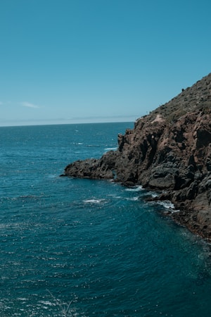 A rugged coastal cliffside meets the expansive ocean, with waves gently lapping against the rocky shore. The sky is clear with a vibrant blue hue, blending with the deep blue of the sea.