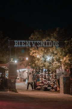An entrance with a large 'Bienvenidos' sign illuminated by warm lights. Underneath, there is a market stall displaying a variety of hats. Trees with hanging lights create a welcoming ambiance. A person is walking towards the stall, adding life to the scene.