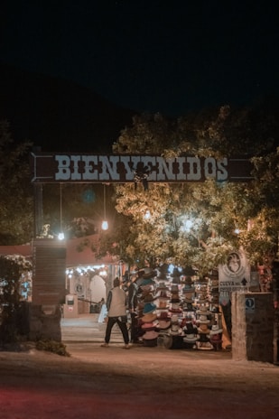 An entrance with a large 'Bienvenidos' sign illuminated by warm lights. Underneath, there is a market stall displaying a variety of hats. Trees with hanging lights create a welcoming ambiance. A person is walking towards the stall, adding life to the scene.