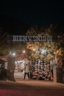 An entrance with a large 'Bienvenidos' sign illuminated by warm lights. Underneath, there is a market stall displaying a variety of hats. Trees with hanging lights create a welcoming ambiance. A person is walking towards the stall, adding life to the scene.