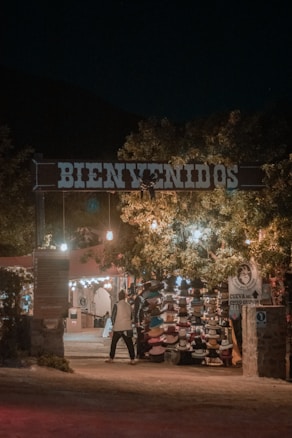 An entrance with a large 'Bienvenidos' sign illuminated by warm lights. Underneath, there is a market stall displaying a variety of hats. Trees with hanging lights create a welcoming ambiance. A person is walking towards the stall, adding life to the scene.