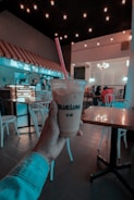 A hand holding a plastic cup with a red straw, featuring the branding 'BLUE LUNA'. The setting appears to be a café with dim lighting and decorative string lights on the ceiling. There are people sitting at tables in the background, with chairs and tables scattered around.