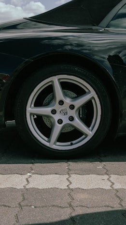 A close-up view of a car's rear wheel with a five-spoke alloy rim and a performance tire. The car's body is sleek and black with a reflective surface. The setting appears to be a paved parking area with interlocking stone tiles.
