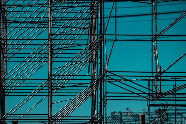 An intricate metal scaffolding structure composed of multiple intersecting bars and beams with a clear blue sky as the background.