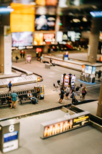 A busy airport baggage claim area with people waiting around. Several baggage carousels can be seen, with people walking or standing nearby. The area is well-lit with colorful advertisements illuminating the scene.