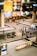 A busy airport baggage claim area with people waiting around. Several baggage carousels can be seen, with people walking or standing nearby. The area is well-lit with colorful advertisements illuminating the scene.