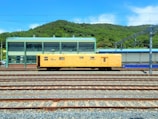 A yellow maintenance train car is positioned on multiple railway tracks in a train yard. In the background, there is a green industrial building with large windows and a green hilly landscape behind it under a clear blue sky.