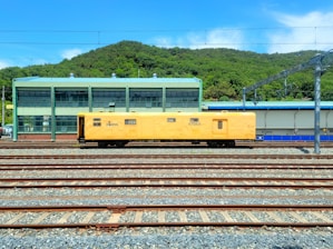 A yellow maintenance train car is positioned on multiple railway tracks in a train yard. In the background, there is a green industrial building with large windows and a green hilly landscape behind it under a clear blue sky.