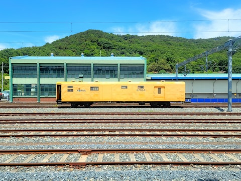 A yellow maintenance train car is positioned on multiple railway tracks in a train yard. In the background, there is a green industrial building with large windows and a green hilly landscape behind it under a clear blue sky.