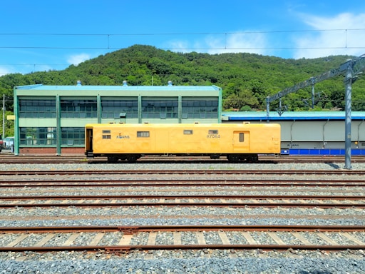 A yellow maintenance train car is positioned on multiple railway tracks in a train yard. In the background, there is a green industrial building with large windows and a green hilly landscape behind it under a clear blue sky.