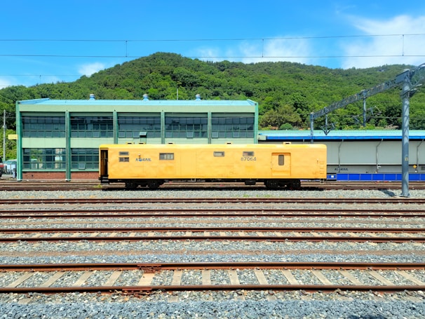 A yellow maintenance train car is positioned on multiple railway tracks in a train yard. In the background, there is a green industrial building with large windows and a green hilly landscape behind it under a clear blue sky.