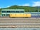 A yellow maintenance train car is positioned on multiple railway tracks in a train yard. In the background, there is a green industrial building with large windows and a green hilly landscape behind it under a clear blue sky.