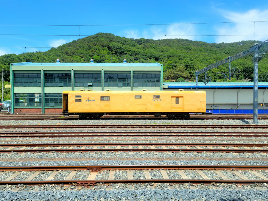 A yellow track maintenance vehicle on multiple railway tracks in a rail yard, with an industrial building behind it.