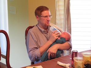 A person is sitting at a dining table, holding a baby wrapped in a blue blanket and feeding them with a bottle. The setting appears to be indoors, with sunlight filtering in through covered windows. Nearby, there are a couple of golden decorative jars and a can of soda on the table.