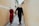 A couple wearing traditional Vietnamese clothing walks hand in hand down a narrow, yellow-walled alley. Both are sporting conical hats known as non la. The woman is dressed in a red ao dai, while the man wears a black one.