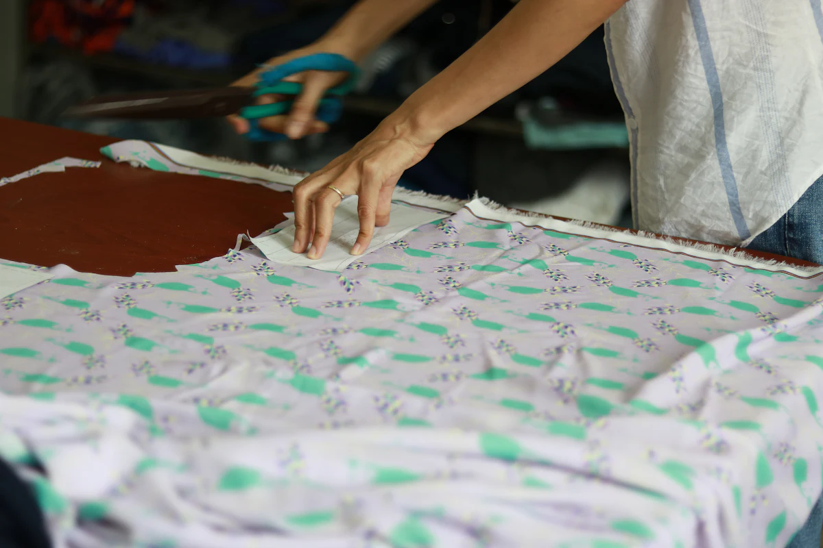 Tangan penjahit menggunting kain bermotif di atas meja kayu, pola kertas tertusuk pin menempel di atas kain, sisa potongan kain berserakan di sisi meja