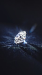 A brilliant cut diamond sits elegantly on a dark surface, reflecting light and casting a delicate play of shadows and sparkles around it.