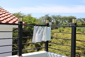 A rooftop terrace with a tiled roof section on the left and a modern railing along the edge. Two cloths are hanging to dry with colorful clips attached to the railing. Lush greenery and trees are visible in the background under a clear blue sky.