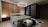 A modern bedroom featuring a minimalist design with a large bed adorned with gray and green bedding. Above the bed hangs an elegant chandelier with crystal-like elements. The room has a warm wooden accent wall and dark, sleek wardrobes on the side. A small bedside table holds a spherical lamp, contributing to the cozy ambiance. The flooring is wooden, complemented by a patterned area rug. A soft glow of light adds a serene atmosphere to the room.