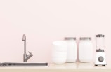 A minimalist kitchen countertop featuring a sleek metal faucet over a built-in sink, a stack of white bowls, two glass jars with metal lids, and a modern white blender with a black base and dial. The backdrop is a soft, light color giving a clean and organized aesthetic.