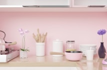 A neatly organized kitchen counter with a pink color theme featuring a small potted plant, a set of wooden cooking utensils in a white container, a white canister, a clear glass jar with a yellow substance, a stack of white plates, a pink saucepan, a purple vase with a purple flower, a white cup, and a modern silver kettle with a pink handle on the stove.