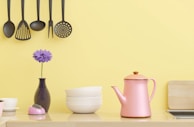 A kitchen scene with hanging black kitchen utensils against a yellow wall. Below, there's a pink kettle, a small stack of white bowls, a purple flower in a vase, and a small pink kitchen timer. A wooden chopping board is partially visible on the right side.