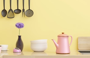 A kitchen scene with hanging black kitchen utensils against a yellow wall. Below, there's a pink kettle, a small stack of white bowls, a purple flower in a vase, and a small pink kitchen timer. A wooden chopping board is partially visible on the right side.