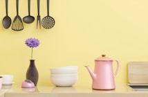 A kitchen scene with hanging black kitchen utensils against a yellow wall. Below, there's a pink kettle, a small stack of white bowls, a purple flower in a vase, and a small pink kitchen timer. A wooden chopping board is partially visible on the right side.