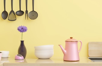 A kitchen scene with hanging black kitchen utensils against a yellow wall. Below, there's a pink kettle, a small stack of white bowls, a purple flower in a vase, and a small pink kitchen timer. A wooden chopping board is partially visible on the right side.