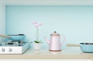 A clean, minimalist kitchen scene featuring a light blue background. On a white countertop, there is a two-burner gas stove with a blue frying pan on the left side, a small potted plant with pink flowers in the middle, a pink teapot with golden accents next to the plant, and a small blue saucepan with a beige handle on the right.