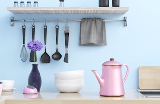 A kitchen scene featuring a light wooden shelf with spice shakers on top, and a metal rod underneath holding various cooking utensils, including a whisk, spoons, and a spatula. A gray towel hangs at one end. A purple flower stands elegantly in a dark vase beside stacked white bowls. A pink teapot with a golden accent sits prominently on the countertop, creating a pastel aesthetic against the light blue wall.