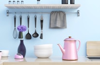 A kitchen scene featuring a light wooden shelf with spice shakers on top, and a metal rod underneath holding various cooking utensils, including a whisk, spoons, and a spatula. A gray towel hangs at one end. A purple flower stands elegantly in a dark vase beside stacked white bowls. A pink teapot with a golden accent sits prominently on the countertop, creating a pastel aesthetic against the light blue wall.