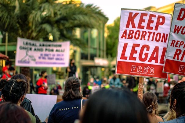 A crowd of people are gathered, holding signs advocating for the legalization of abortion. Prominent signs display messages such as 'Keep Abortion Legal.' The setting appears to be an outdoor protest or demonstration, with palm trees and a building in the background.