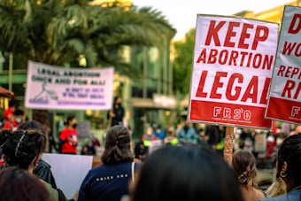 A crowd of people are gathered, holding signs advocating for the legalization of abortion. Prominent signs display messages such as 'Keep Abortion Legal.' The setting appears to be an outdoor protest or demonstration, with palm trees and a building in the background.