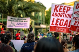 A crowd of people are gathered, holding signs advocating for the legalization of abortion. Prominent signs display messages such as 'Keep Abortion Legal.' The setting appears to be an outdoor protest or demonstration, with palm trees and a building in the background.