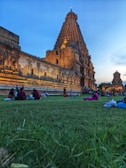 A large, intricately carved temple made of stone stands under a clear sky with a golden hue from the setting sun. People are seen sitting on a lush green lawn in front of the temple, adding a lively atmosphere to the historical setting.
