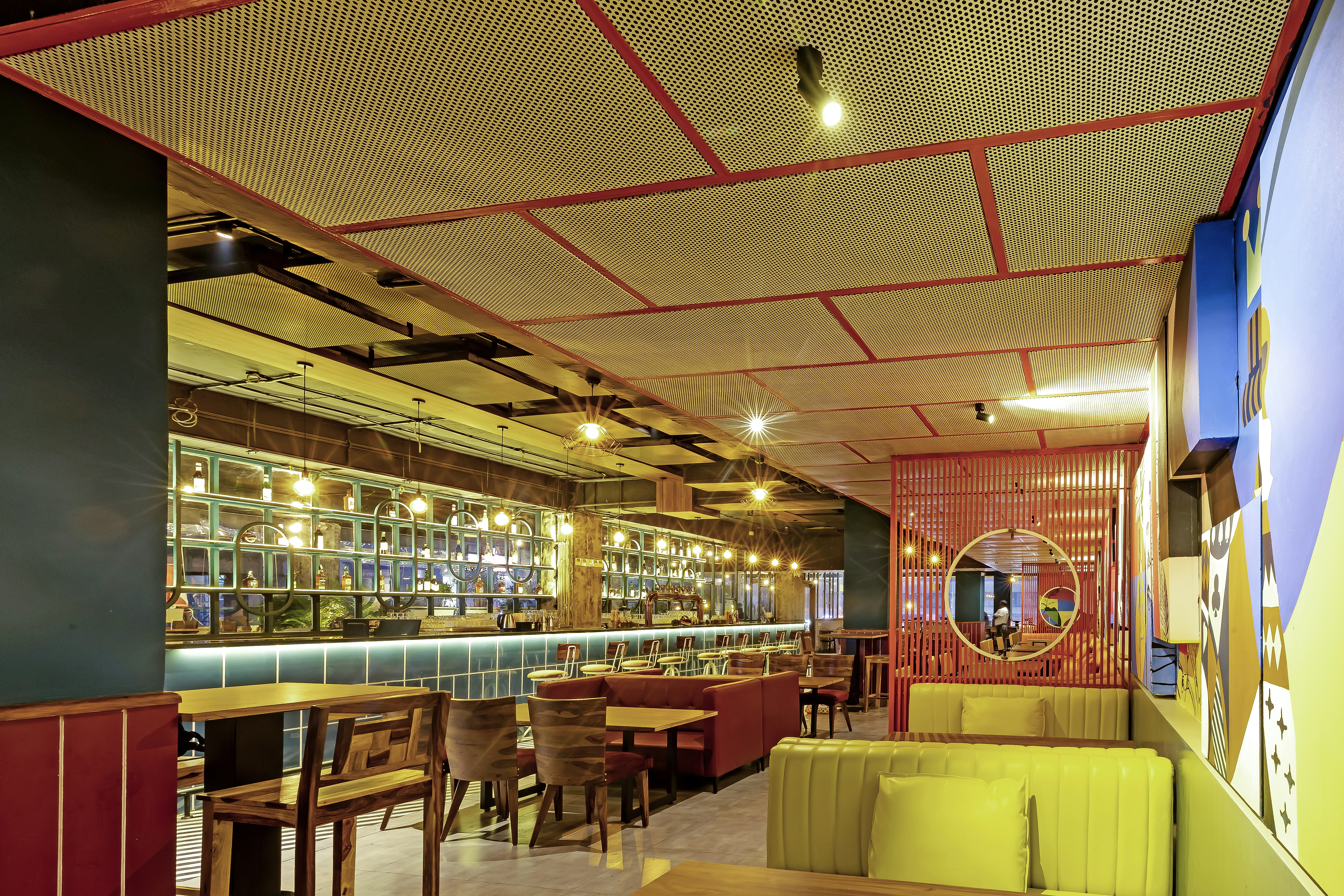 An interior view of a modern, well-lit restaurant with a mix of wooden and upholstered furniture. The ceiling has a grid-like pattern with integrated lights. The walls are decorated with mirrors, glass panels, and abstract art. The seating arrangement includes wooden chairs, cushioned benches, and red booths. A circular mirror is mounted on a red lattice wall, adding a touch of contemporary design.