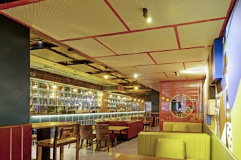 An interior view of a modern, well-lit restaurant with a mix of wooden and upholstered furniture. The ceiling has a grid-like pattern with integrated lights. The walls are decorated with mirrors, glass panels, and abstract art. The seating arrangement includes wooden chairs, cushioned benches, and red booths. A circular mirror is mounted on a red lattice wall, adding a touch of contemporary design.