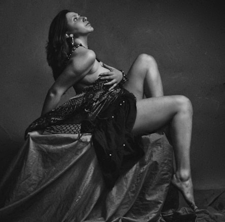 A person poses artistically while leaning back against a fabric-covered structure. Their posture is dramatic and expressive, with elegant drapery adding to the aesthetic appeal. The black and white filter enhances the contrast and mood.