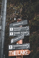 Several directional signs are mounted on a wooden post, pointing towards various destinations including hiking trails and a city center. The signs have text in English and another language, with small symbols indicating activities and distances. The background features dense, dark green foliage, suggesting a forested area.
