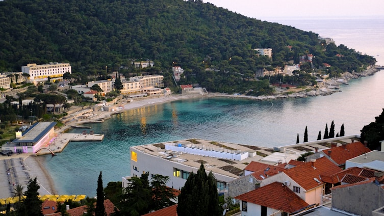 A coastal landscape features a bay with turquoise water, surrounded by lush green hills. Several buildings line the water's edge, including a hotel and a cluster of orange-roofed houses. Sandy beaches are scattered along the shore, populated with a few people. Vegetation, such as pine trees and shrubs, cover the hillside and the surrounding areas.