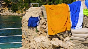 Colorful towels and swim trunks hang on a rocky wall by a clear, blue body of water, with lush greenery and a rocky landscape in the background.