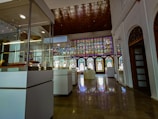 An elegant interior space with glass display cases containing various artifacts. A large stained glass window stretches across one side of the room, casting colorful light onto the polished floor. The ceiling features intricate wooden paneling, and white walls with decorative arches add to the historical ambiance.