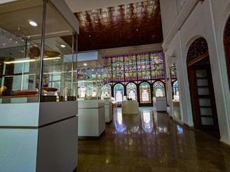 An elegant interior space with glass display cases containing various artifacts. A large stained glass window stretches across one side of the room, casting colorful light onto the polished floor. The ceiling features intricate wooden paneling, and white walls with decorative arches add to the historical ambiance.