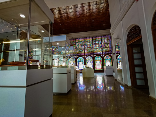 An elegant interior space with glass display cases containing various artifacts. A large stained glass window stretches across one side of the room, casting colorful light onto the polished floor. The ceiling features intricate wooden paneling, and white walls with decorative arches add to the historical ambiance.